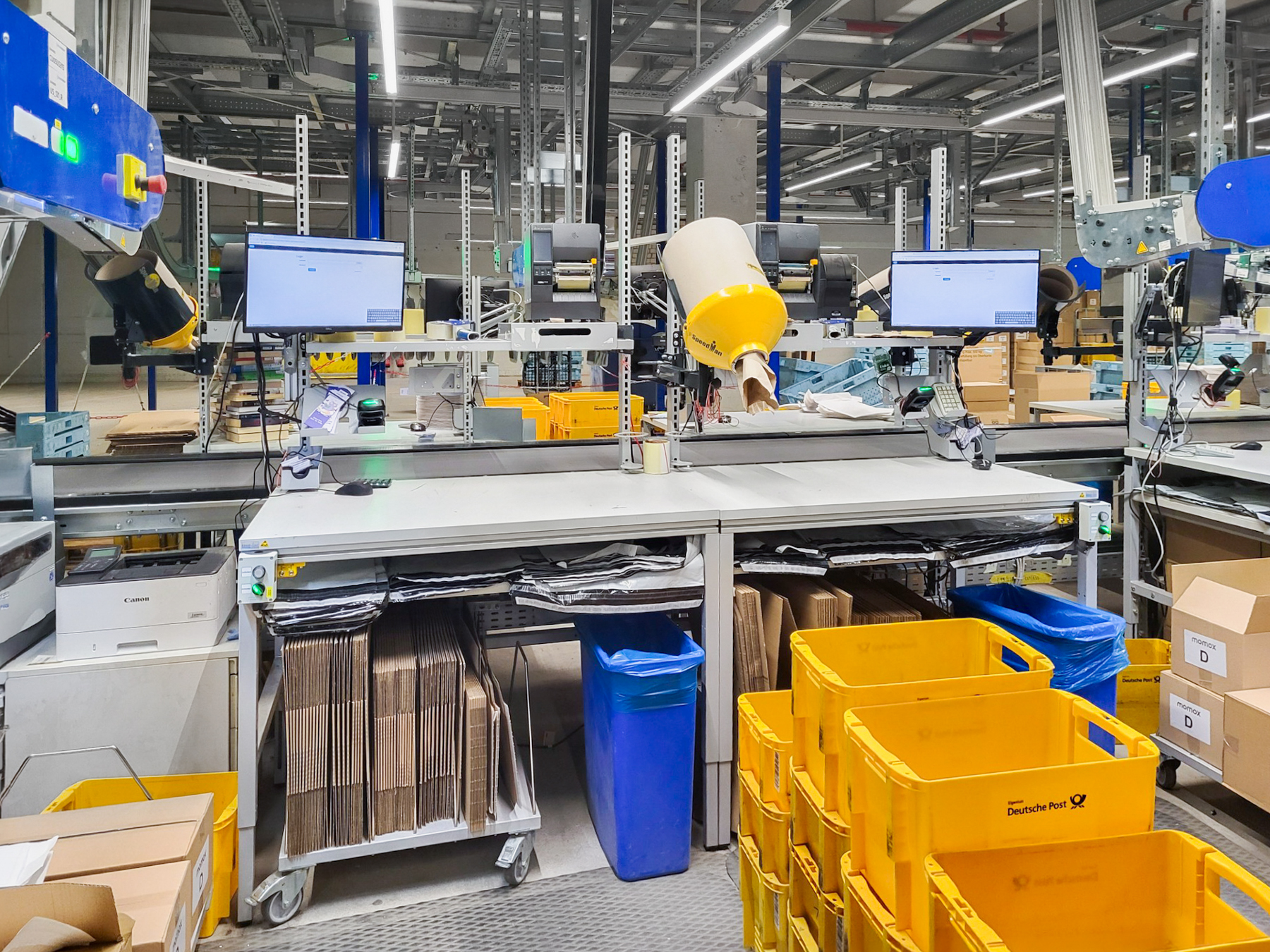 ERGO-line-Packtisch-ERGO-Basic-momox_Leipzig-004 Table d'emballage ERGO-line dans le secteur logistique, équipée d'écrans, d'un scanner, d'une tablette de rangement, d'un magasin à boîtes pliantes mobile et d'étagères